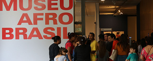 Fotografia retangular. Do lado esquerdo tem um painel e nele está escrito, com destaque e na cor vermelha: “Museu Afro Brasil”. Do lado direito tem várias pessoas que parecem caminhar em direção de uma porta de vidro. Há homens, mulheres e crianças.