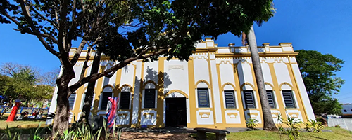 Fotografia retangular de uma edificação antiga, pintada em tons de amarelo. Na frente tem um grande gramado, um banco e uma árvore. Nas laterais da foto também há imagens parciais de outras árvores.