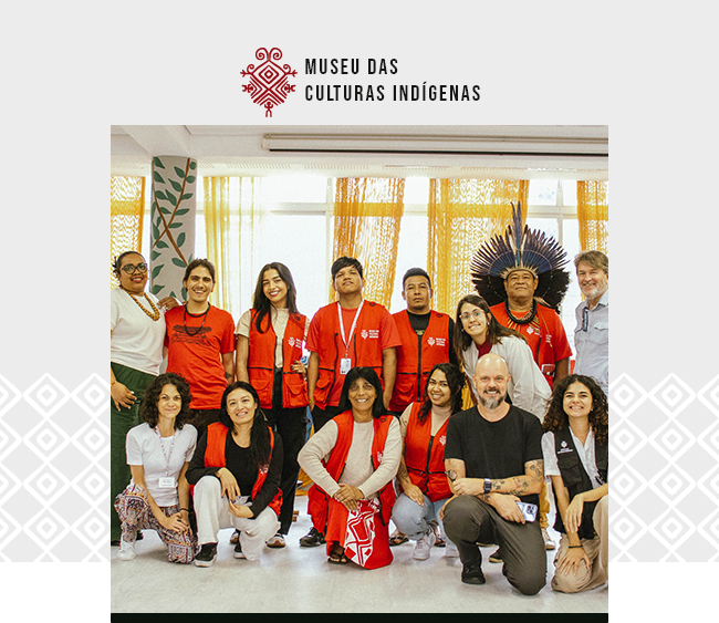 A imagem mostra um grupo diverso de pessoas posando juntas para uma foto em um ambiente interno ao Museu das Culturas Indígenas. O grupo é composto por diversas pessoas, algumas em pé e outras agachadas na frente. A maioria usa coletes vermelhos com o logo do museu. Duas pessoas se destacam: uma delas, ao fundo, usa um grande elemento de penas na cabeça, tradicional da cultura indígena, e veste uma camiseta vermelha. Outra pessoa, um homem com barba e camiseta preta, está agachada na frente do grupo, à direita. As expressões das pessoas são amigáveis e sorridentes. Ao fundo, as janelas estão cobertas por cortinas amarelas, e há elementos de decoração com motivos naturais, como plantas.